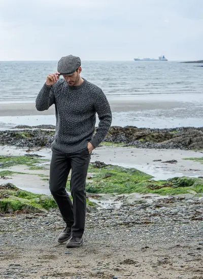 Man Walking on Beach Wearing Paddy Cap and Oisin Aran Crew Neck Sweater in Slate
